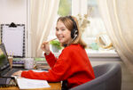 A girl wearing headphones and a red sweater smiles as she learns at-home in her online classroom.